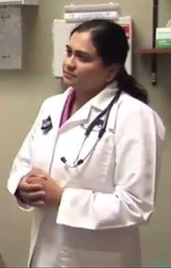 The image shows a woman wearing a white lab coat, standing upright with her hands clasped together, looking directly at the camera.