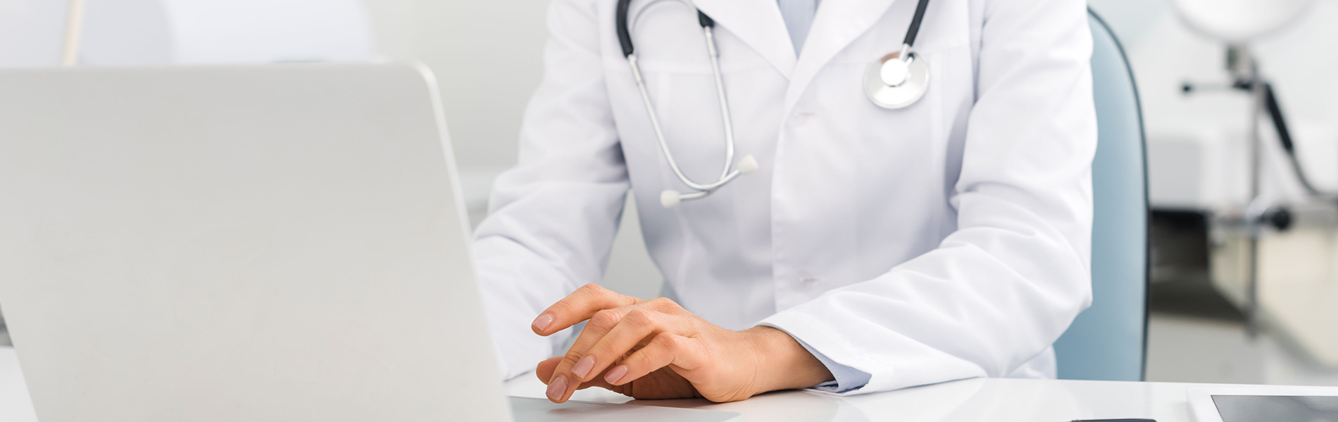 A woman in a white lab coat is sitting at a desk with a laptop, wearing glasses and looking down at the screen.