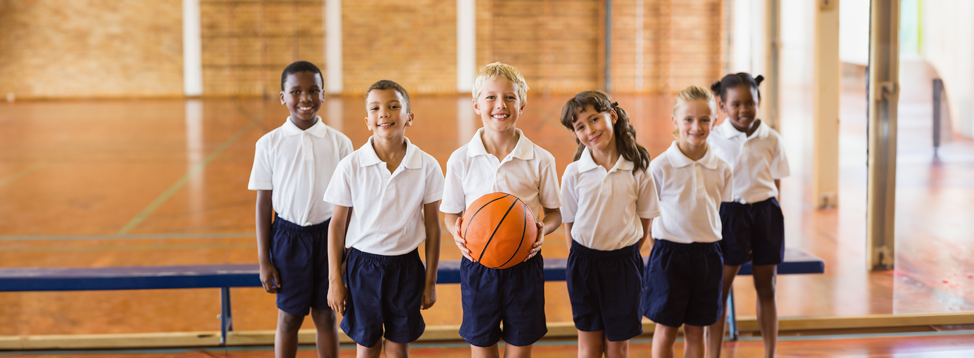 A group of young people wearing school uniforms pose together on a basketball court with benches in the background.