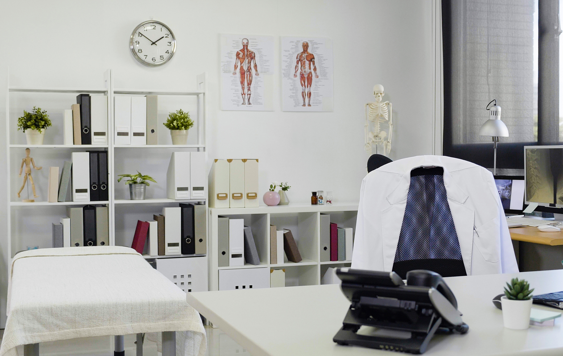 The image shows an office space with a desk, chair, computer monitor, and various items on shelves and a table, including medical models, books, and decorative objects.