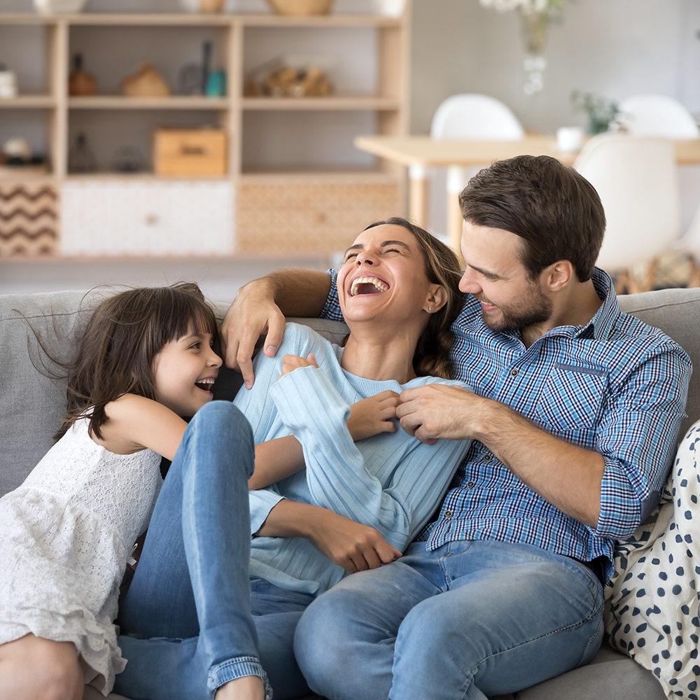 A family of four, including two adults and two children, seated on a couch, sharing a joyful moment with smiles and laughter.