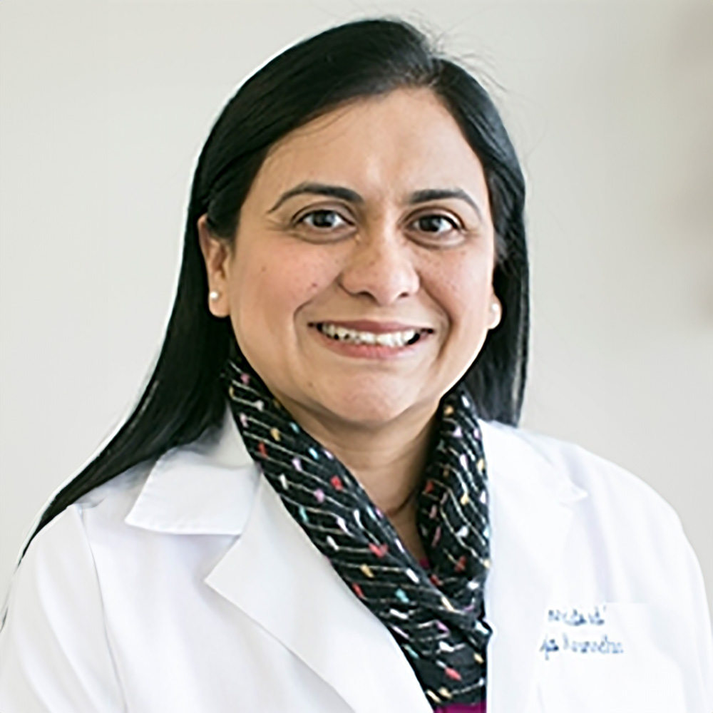 The image shows a woman standing against a light background. She has dark hair, is wearing glasses, and has a smile on her face. Her attire includes a white lab coat with black trim, a patterned scarf, and a pink top underneath. There s a name tag visible on the lab coat, but the text is not legible in the image provided.